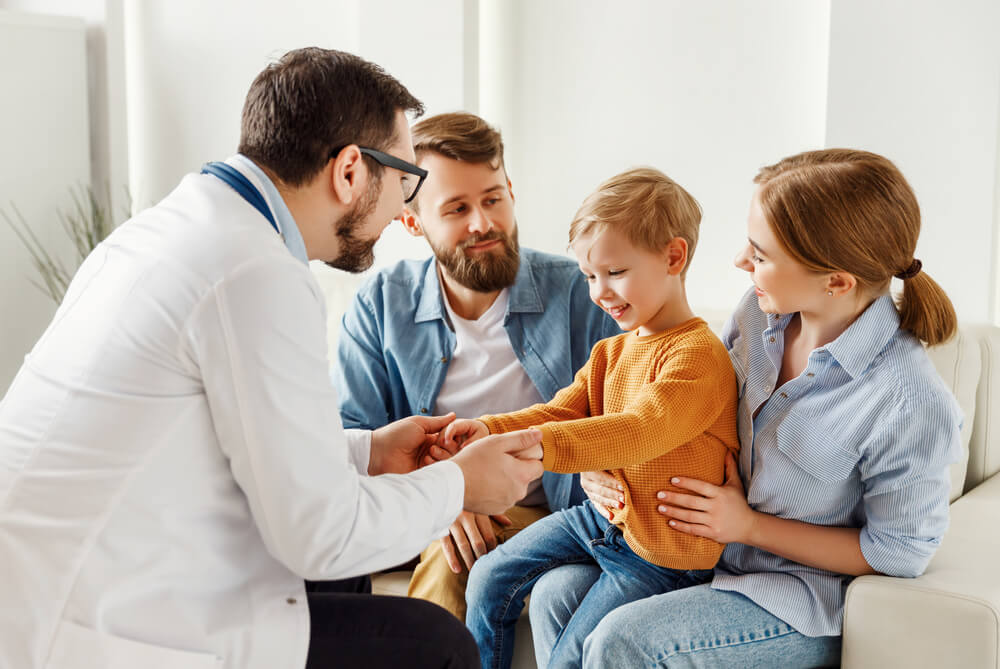 Positive male pediatrician talking with happy boy and parents during check up appointment in cozy office in modern clinic