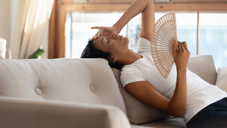 Tired unwell young African American woman sit lie on couch at home use waver suffer from hot weather indoors. Overheated millennial biracial female renter breathe fresh air from hand fan.