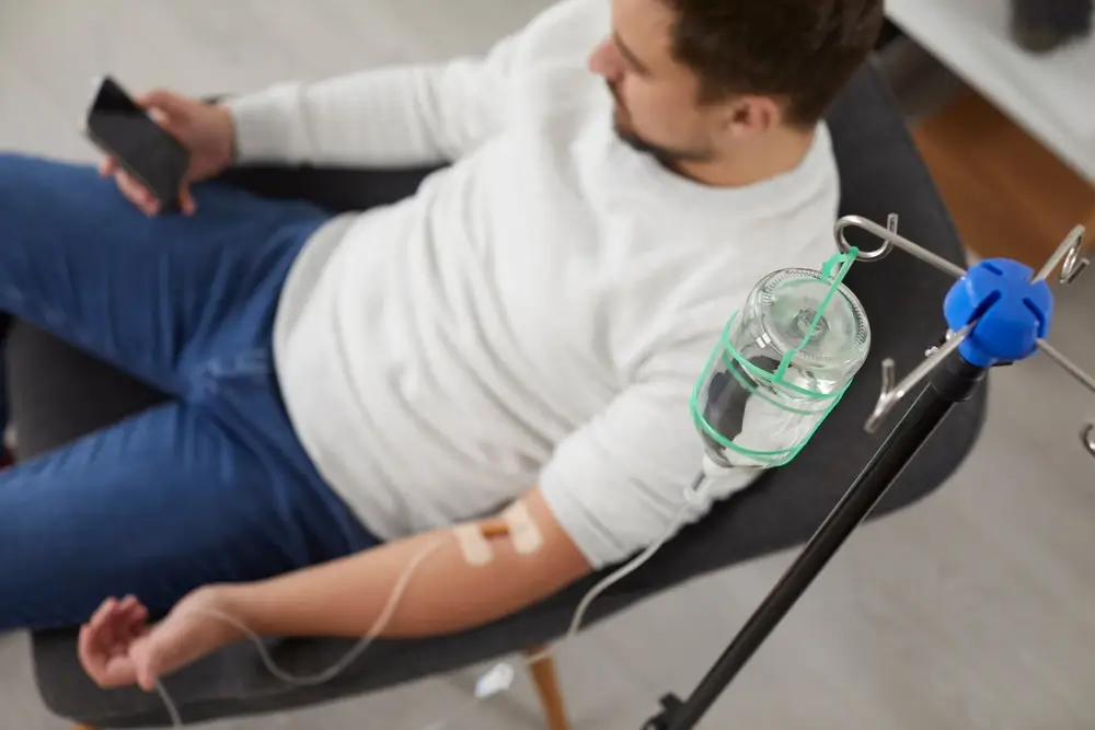 Top view photo of a man sitting in the chair and using the smartphone while receiving IV drip infusion and vitamin therapy in his blood.