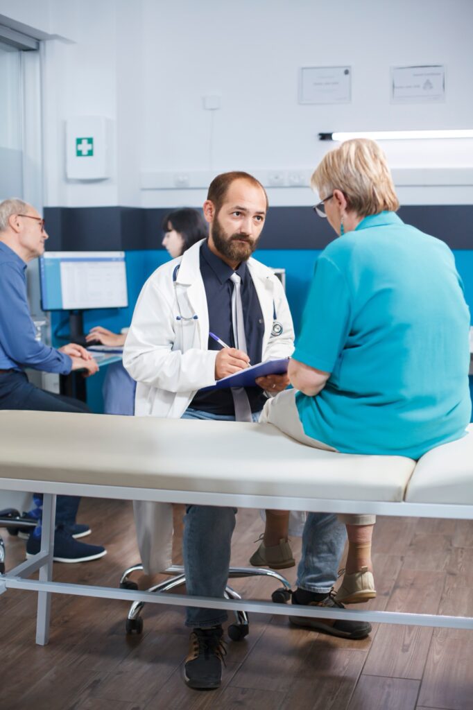 Aged woman with physical injuries receives advice from male practitioner at clinic. Medical assistant examining elderly patient for orthopedic diagnosis and treatment while discussing pain management.