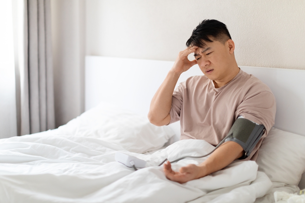 Unhealthy asian man in pajamas sitting in bed at home, checking blood pressure on his own