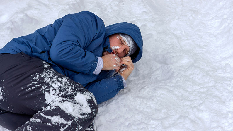 Frozen man in a blue jacket and hat lying down covered snow and frost, trying to stay warm on a very cold winter day, snow falls around him. Sick mountaineer with hypothermia on snow during the day.