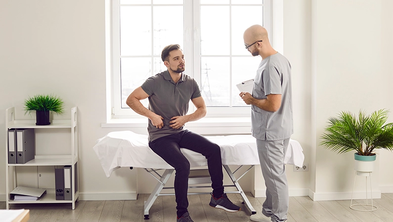 Young man sitting on the couch in the doctor's office and pointing to his stomach to the doctor during medical examination in clinic. Physician listening to the patient's complaints.