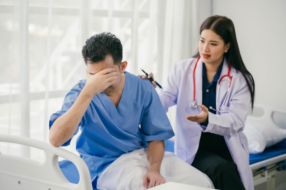 A man in a hospital bed is experiencing migraine while a woman in a white coat stands over him.