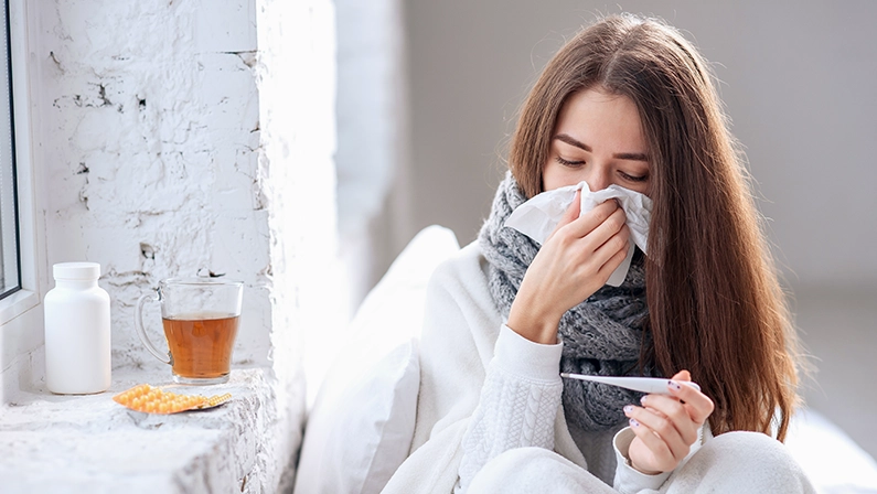 Cold and flu. Sick woman caught cold, feeling illness and sneezing in paper wipe. Closeup of beautiful unhealthy girl covered in blanket wiping nose and looking at thermometer. Healthcare concept.